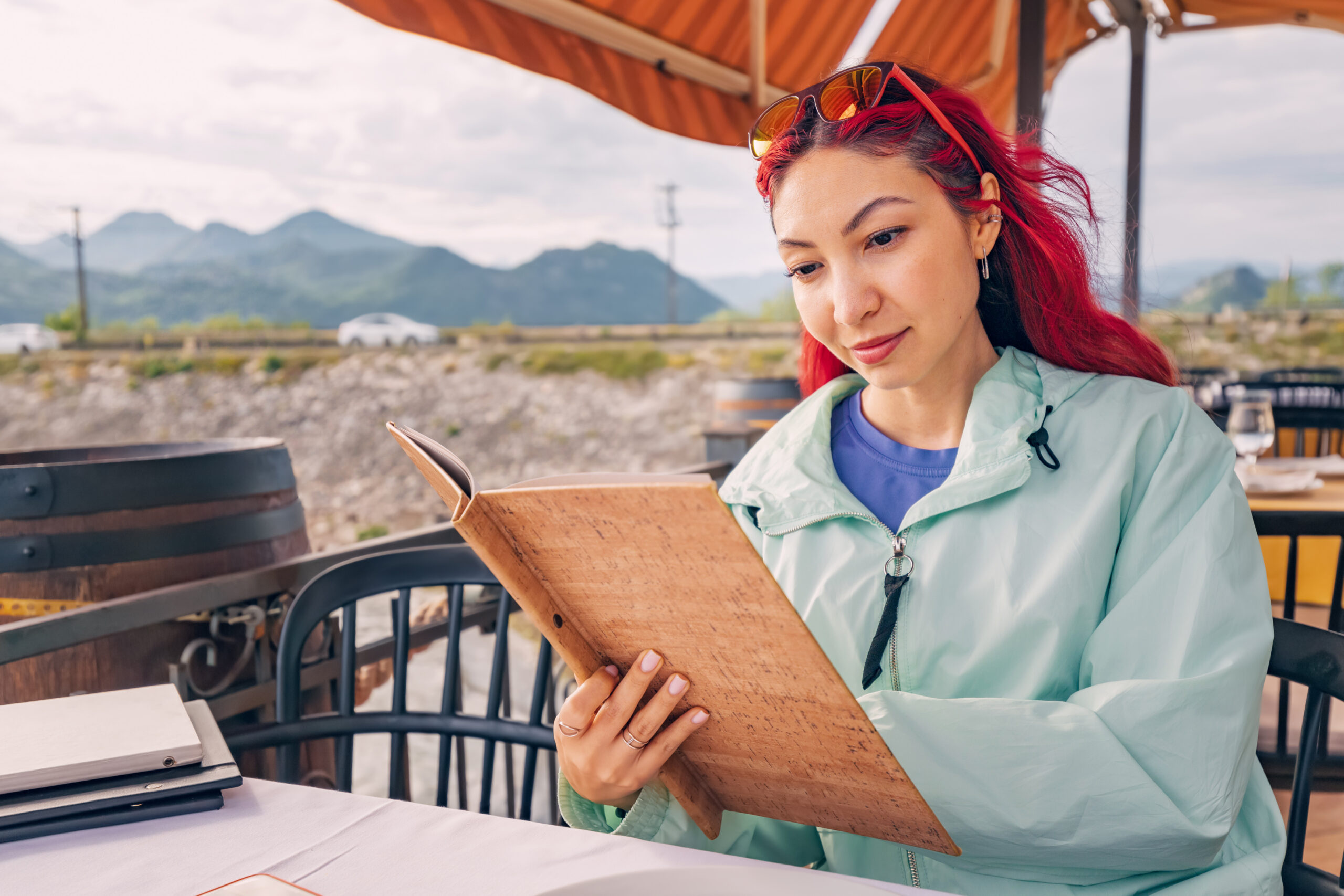 A young woman smiles while reading the menu at a lakeside restaurant, enjoying the panoramic view o...